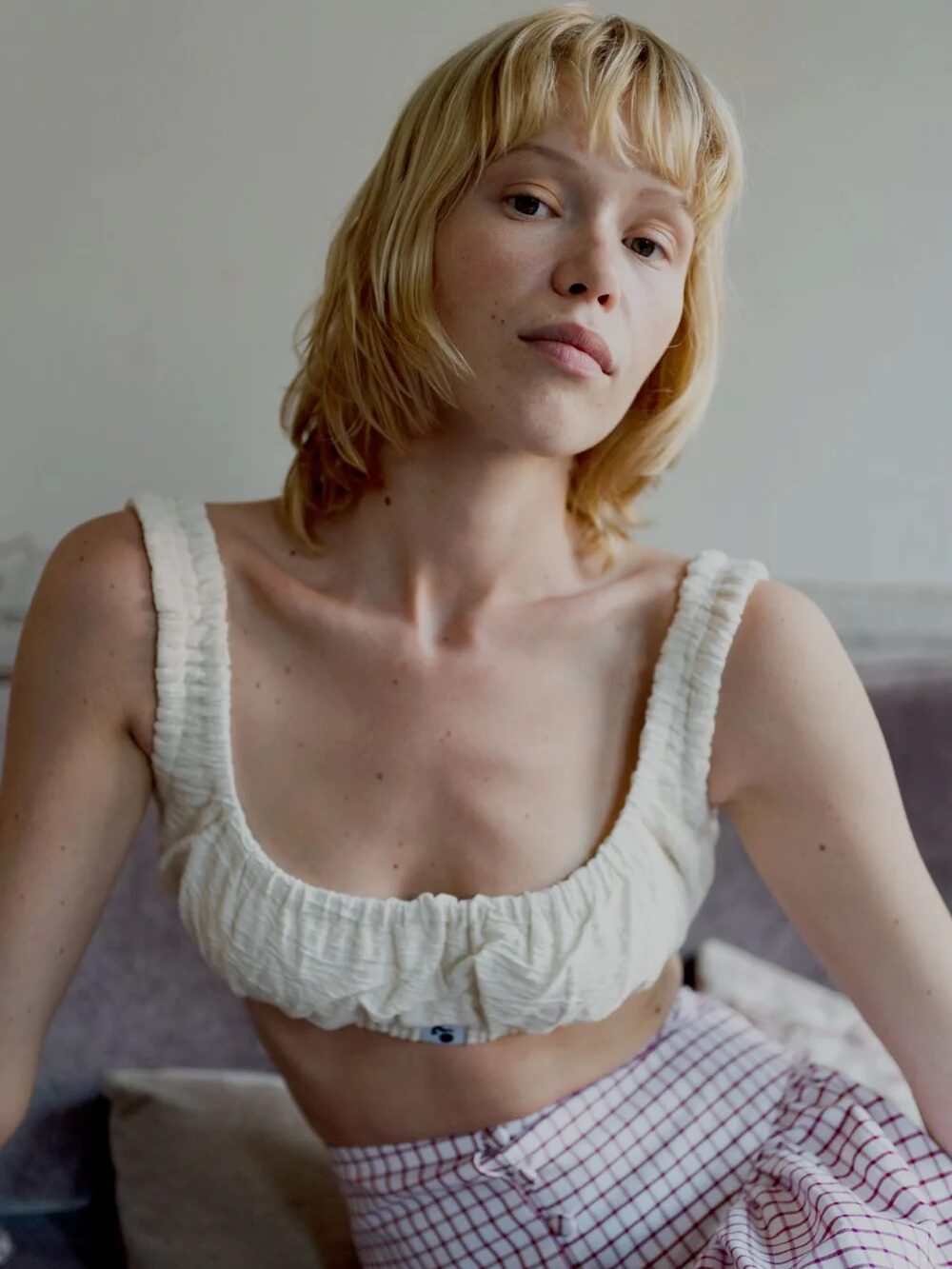 A woman with short blonde hair sits casually, wearing a cream ruffled top and red plaid pants, looking directly at the camera.