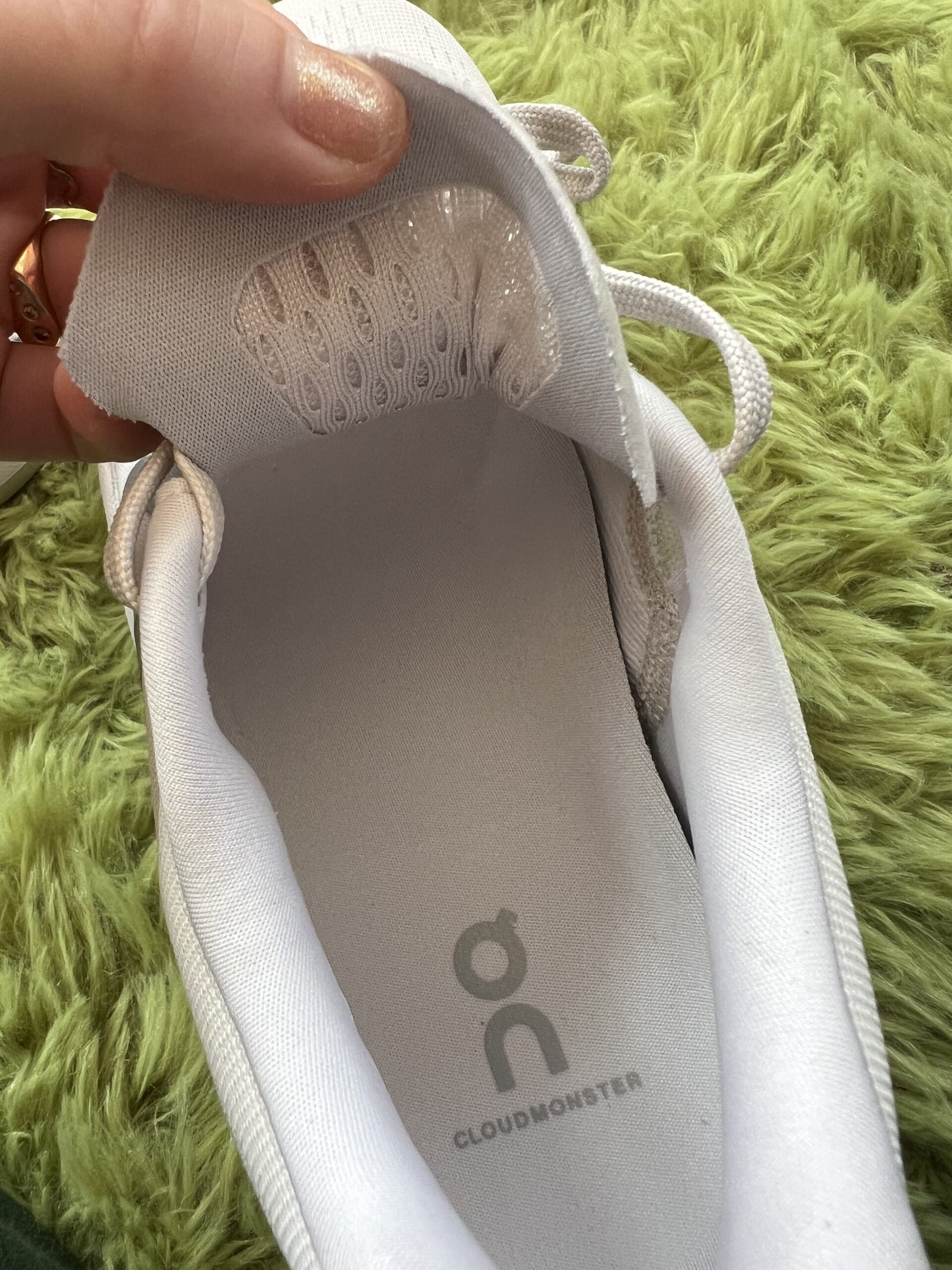 A person's hand holding open a light gray shoe with white shoelaces, showing the inner sole branded "cloudmonster" with a circular logo.