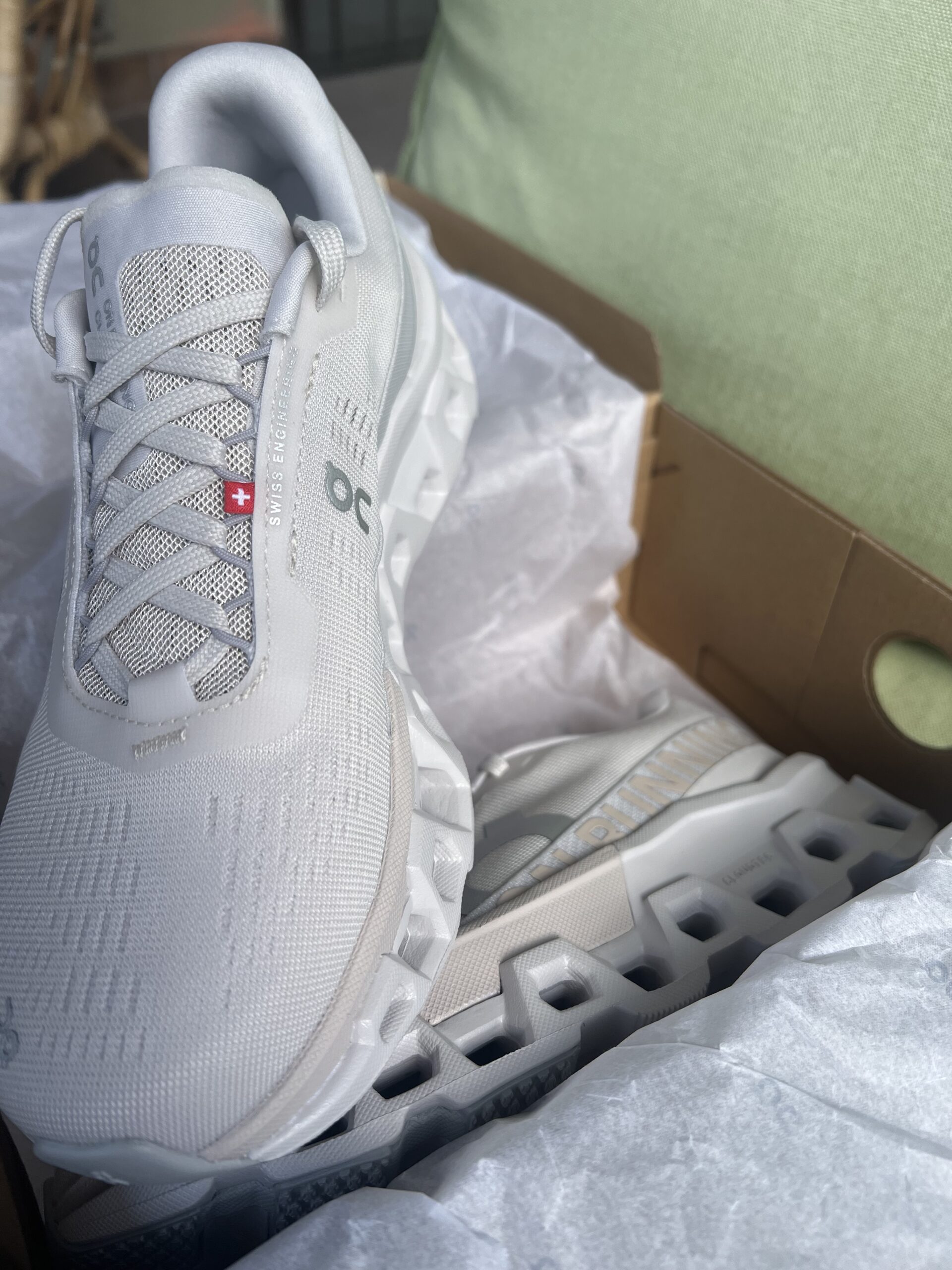 A new white running shoe with distinctive thick soles, displayed on white paper next to a cardboard box and a green pillow.