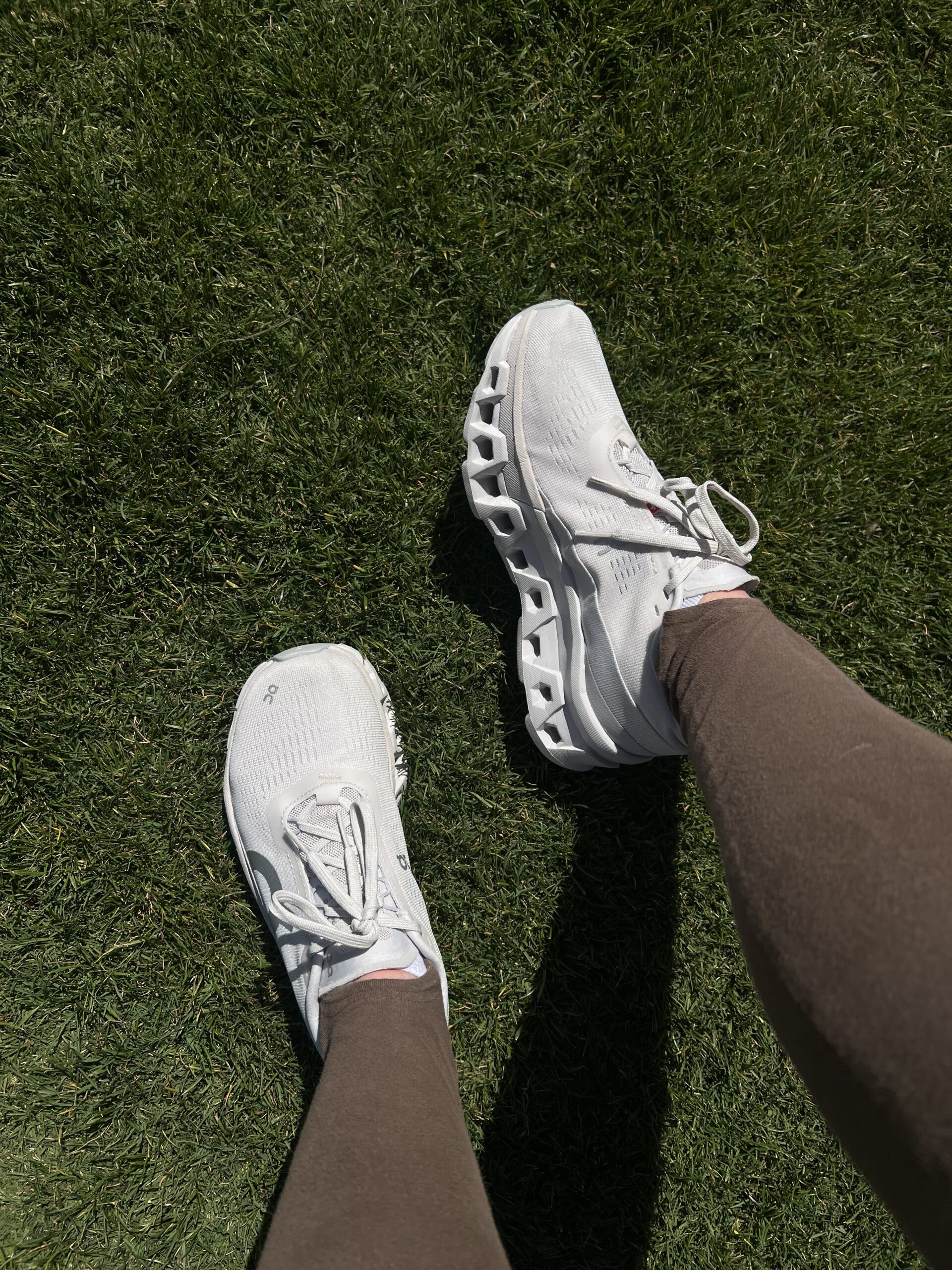 A person wearing white sneakers and brown leggings, standing on lush green grass.