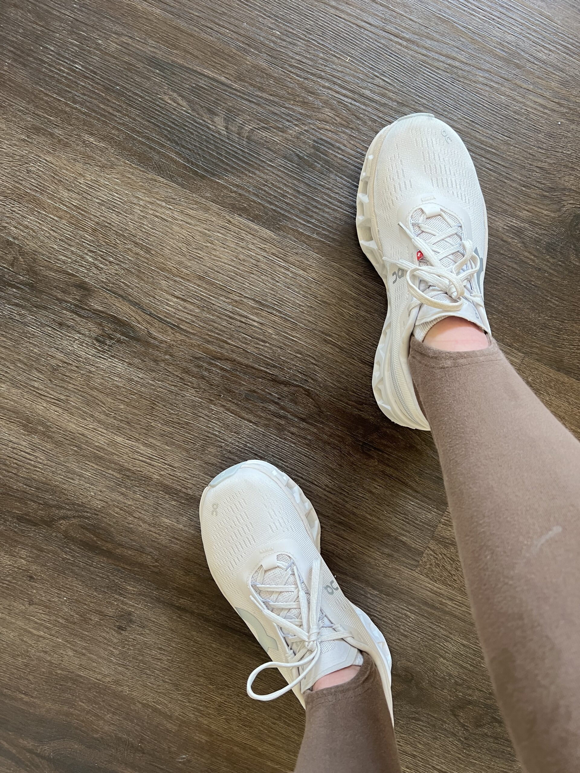 A person wearing white sneakers with visible toes standing on a wooden floor.