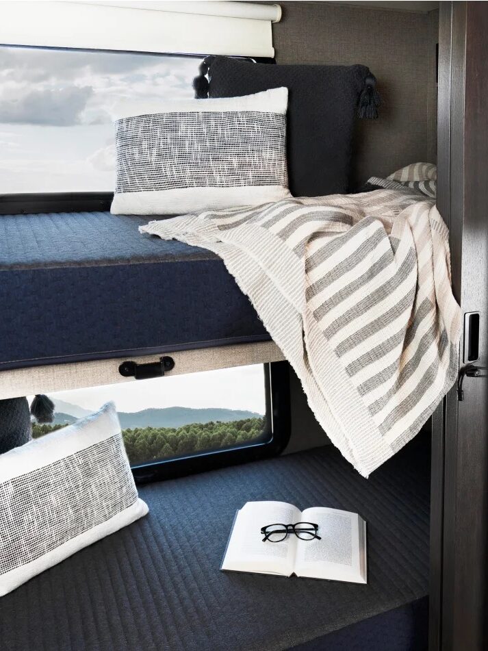 Interior of a camper van showing a cozy bunk bed setup with pillows, a blanket, and a book by the window, overlooking a scenic view.
