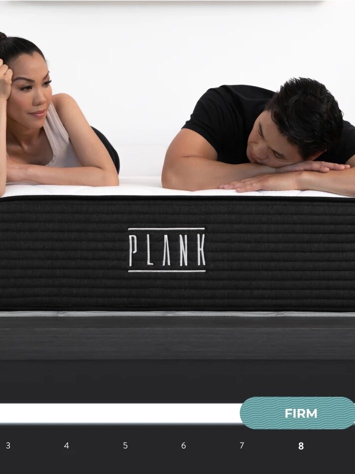A man and woman examine a dual-sided 'plank' firmness mattress displayed on a scale from softest to extra firm.