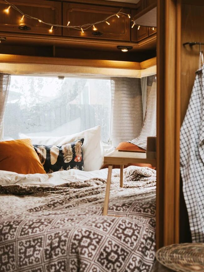 Cozy camper van interior featuring a bed with patterned bedding, fairy lights, curtains, and wooden cabinets, creating a warm and inviting atmosphere.