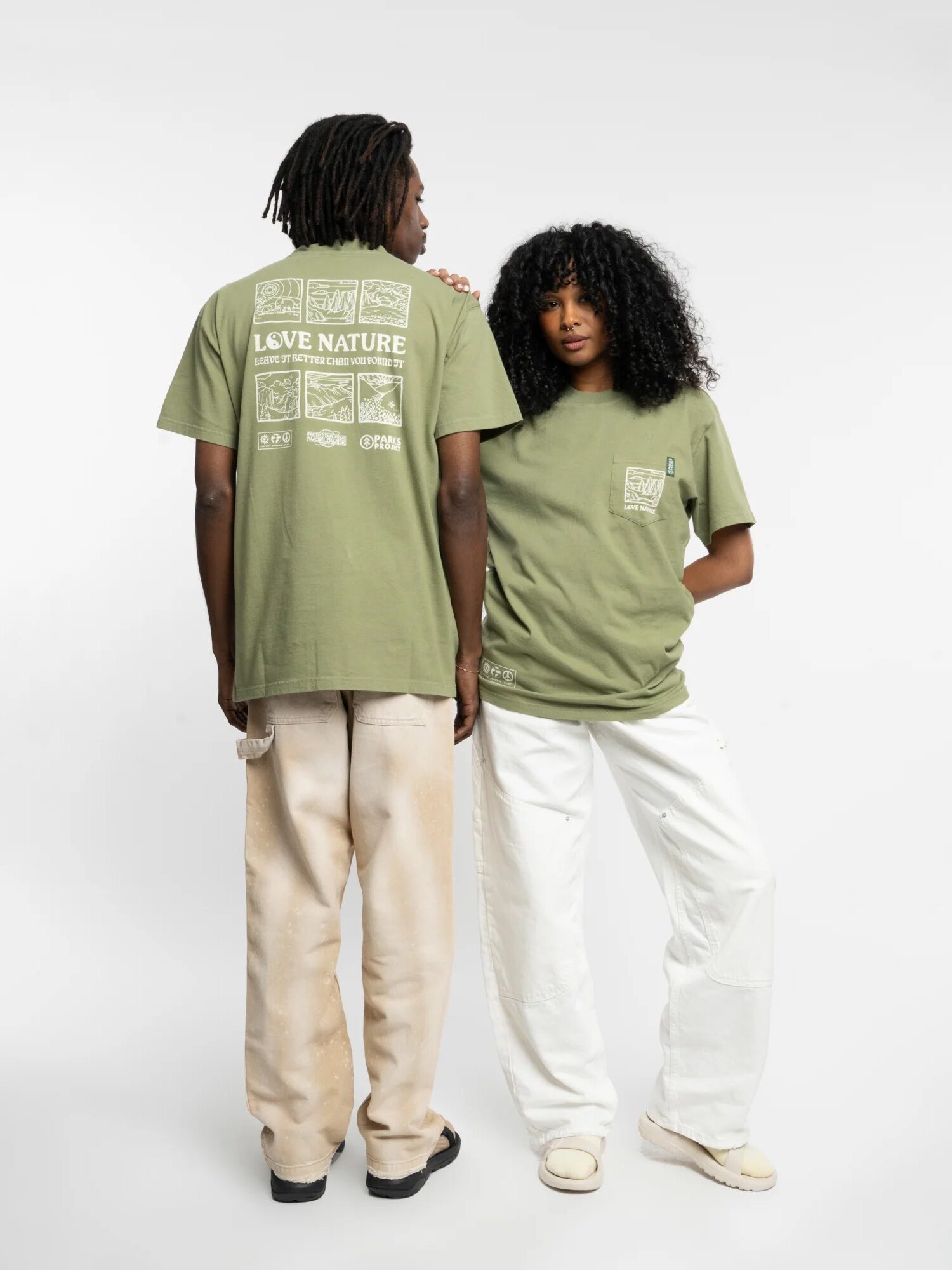 Two people, one man and one woman, stand back-to-back wearing green t-shirts with environmental slogans. the man is in beige pants, the woman in white, both facing away from the camera.