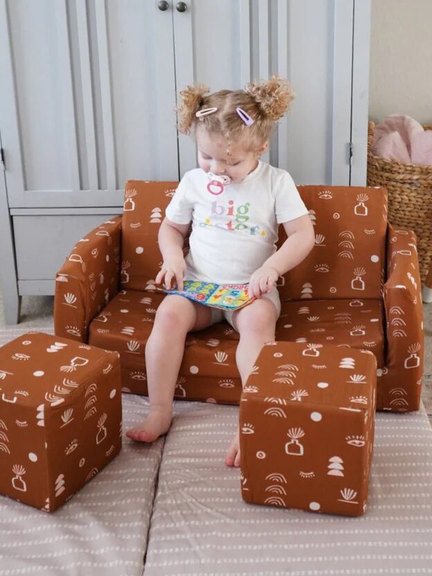 Toddler sitting on a small patterned armchair with matching ottoman, surrounded by child-friendly furniture.