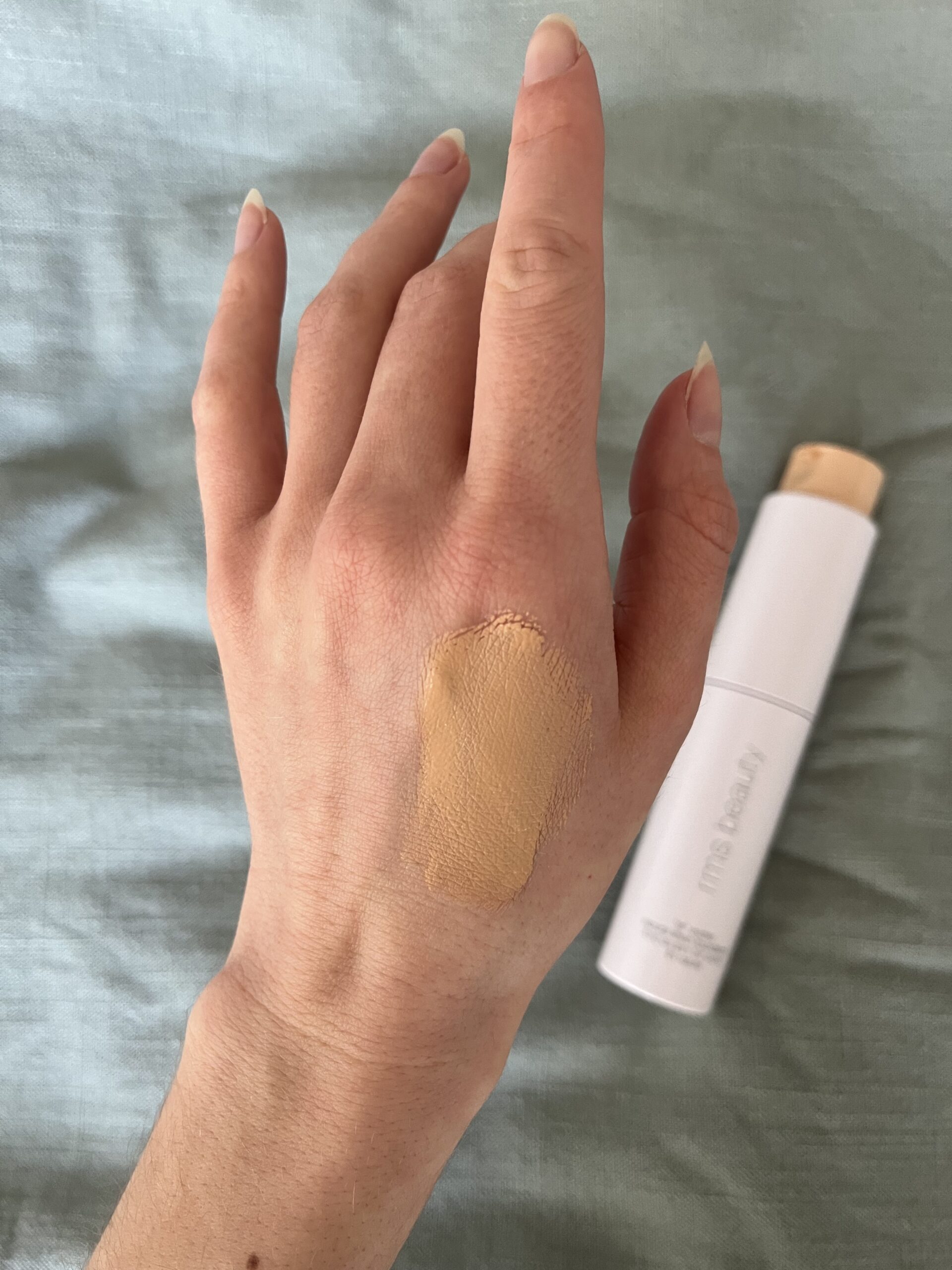 A hand with a swatch of beige foundation on the back, next to a tube of foundation with a white cap.