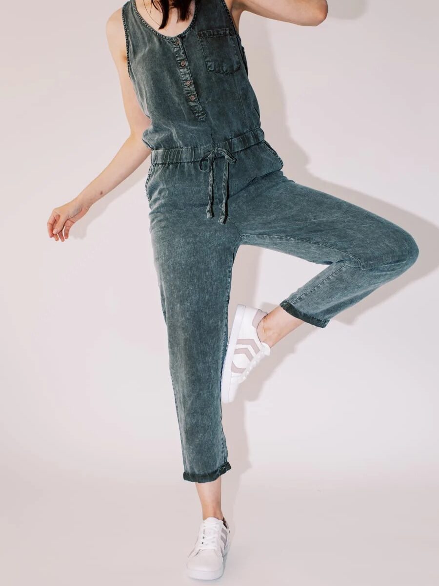 Person in a denim jumpsuit striking a playful pose on one leg against a light background.
