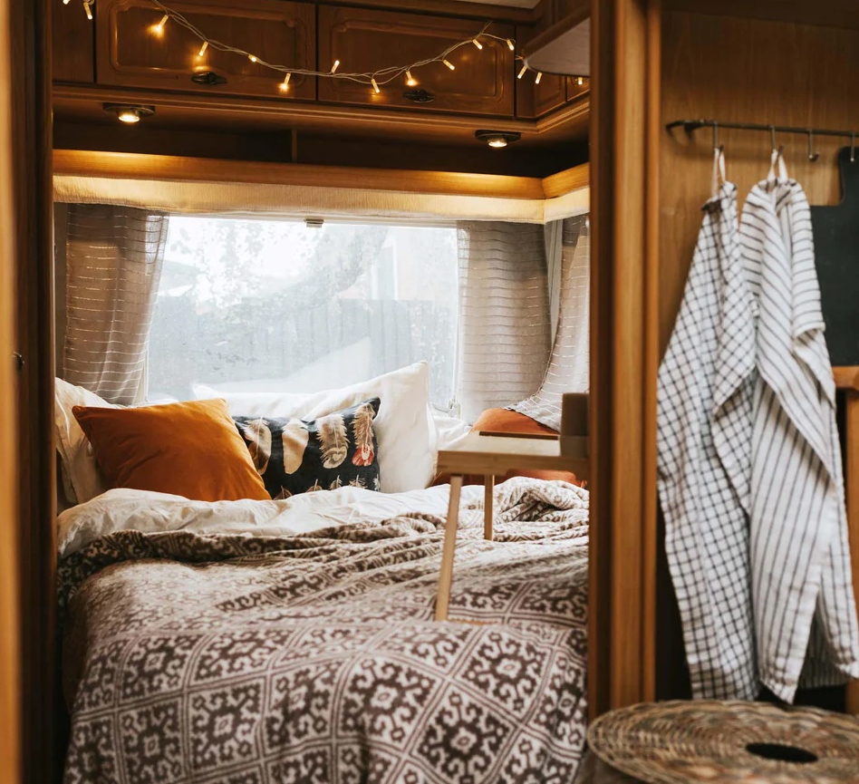 Cozy camper van interior with a bed adorned with patterned bedding and multiple pillows, a small wooden table, hanging clothes, and string lights.