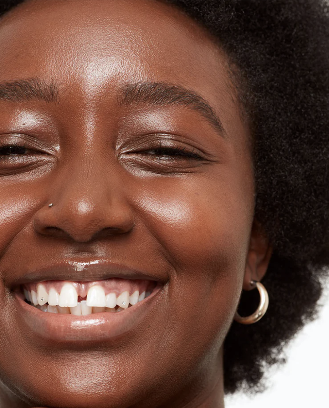 Close-up of a smiling black woman with curly hair, showcasing a small earring and a "new" sticker on her forehead.