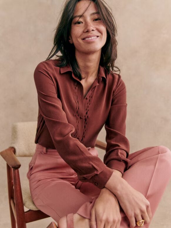 A woman in a rust-colored blouse and pink trousers sitting with one leg crossed over the other, smiling gently at the camera.