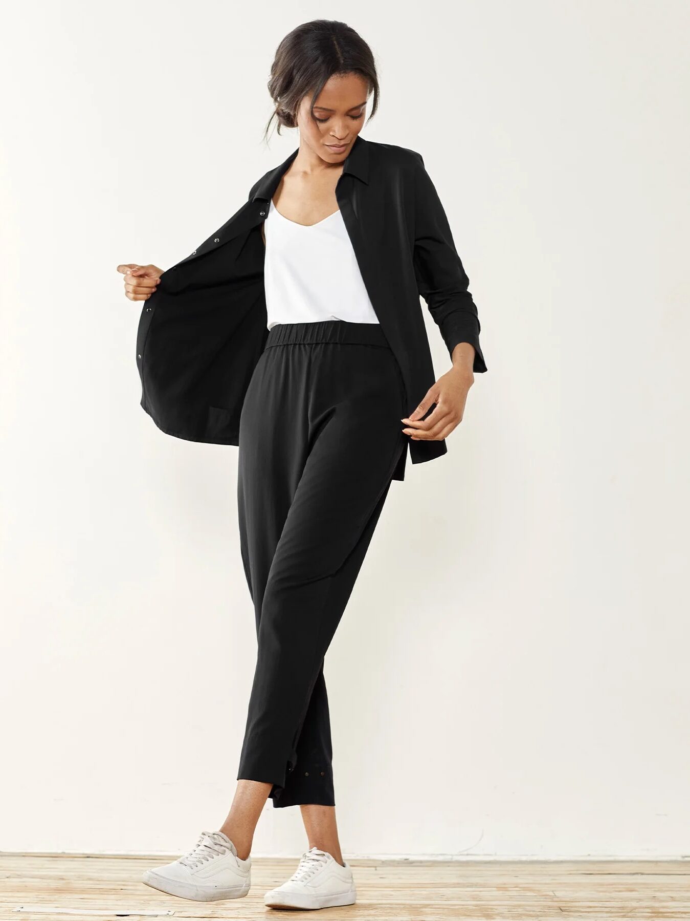A woman in a black blazer and trousers with white sneakers posing against a neutral background.