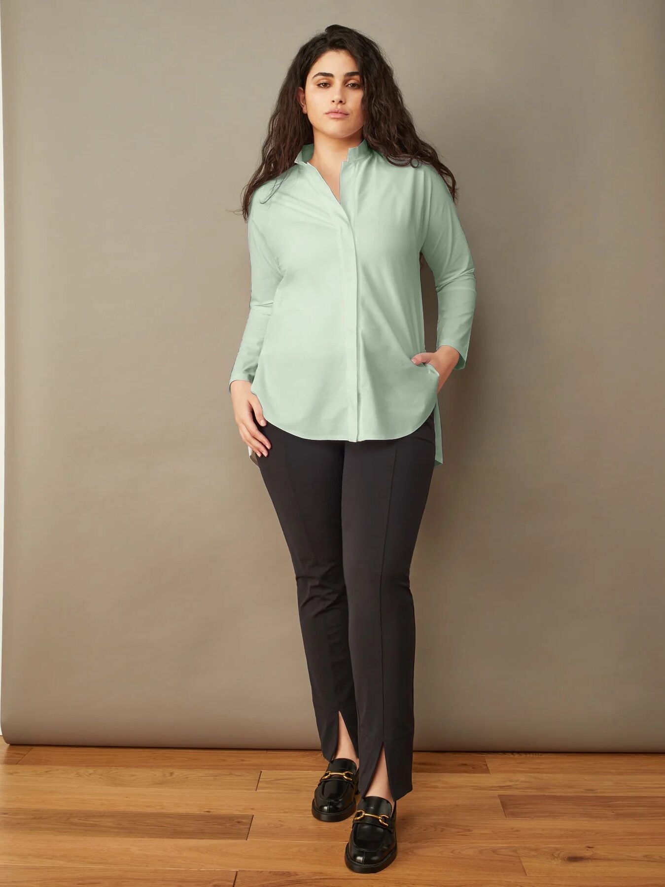 Woman standing against a neutral backdrop, wearing a light green blouse, black trousers, and black shoes.