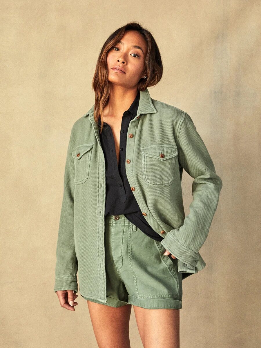 A woman in a casual olive green jacket and shorts stands against a neutral backdrop.