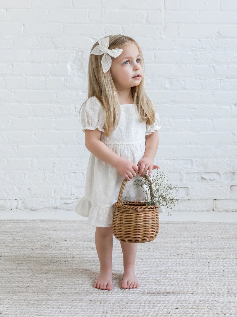 A young girl in a white dress holding a basket stands barefoot, looking away thoughtfully, against a white brick wall.