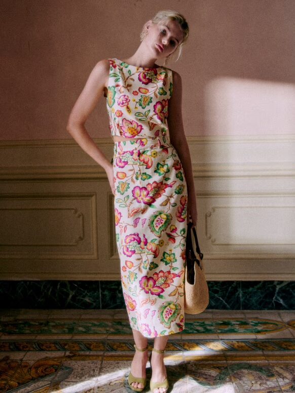 A woman in a floral dress stands in a room with vintage decor, holding a straw bag and wearing green sandals.
