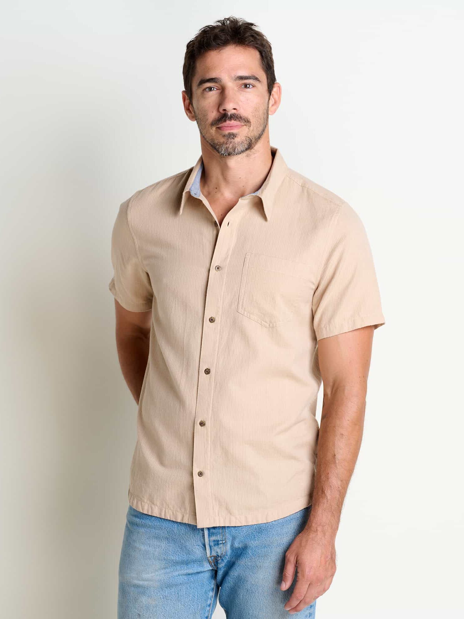 Man standing against a white background, wearing a beige short-sleeve shirt and blue jeans, looking directly at the camera.