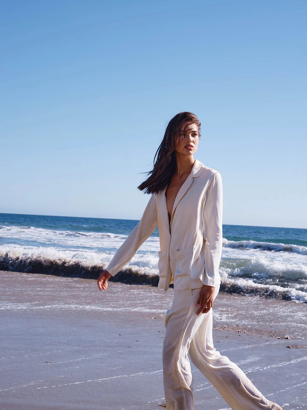 A woman in a white suit walks along a sandy beach with the ocean and waves in the background.