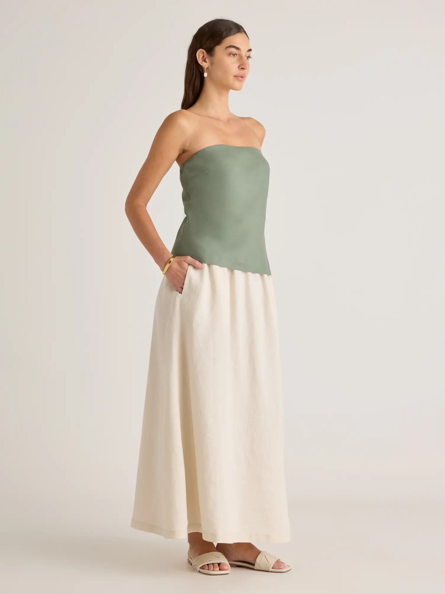 A woman stands in a studio, wearing a strapless green top and cream wide-leg trousers, looking to the side.
