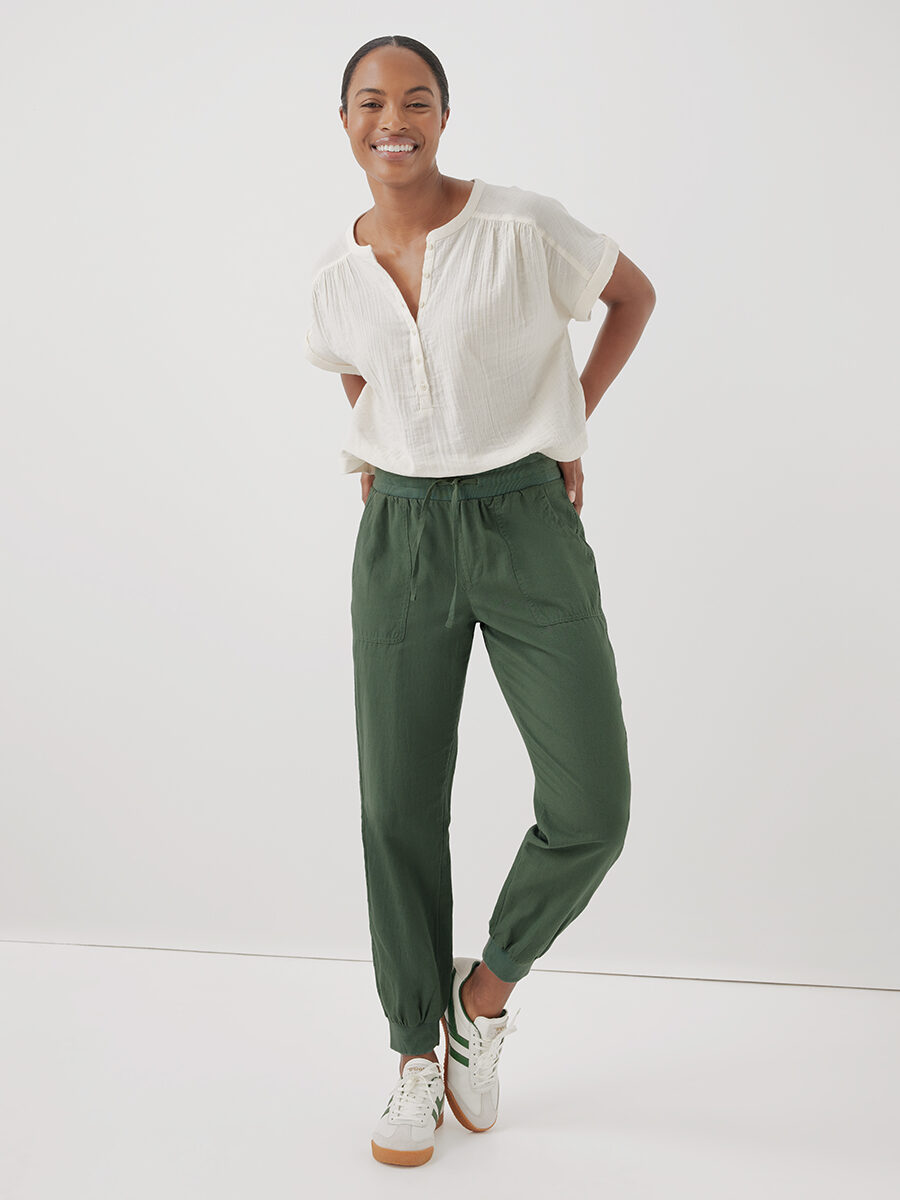 A woman stands smiling, hands in pockets, wearing a white blouse, green trousers, and white sneakers against a plain background.