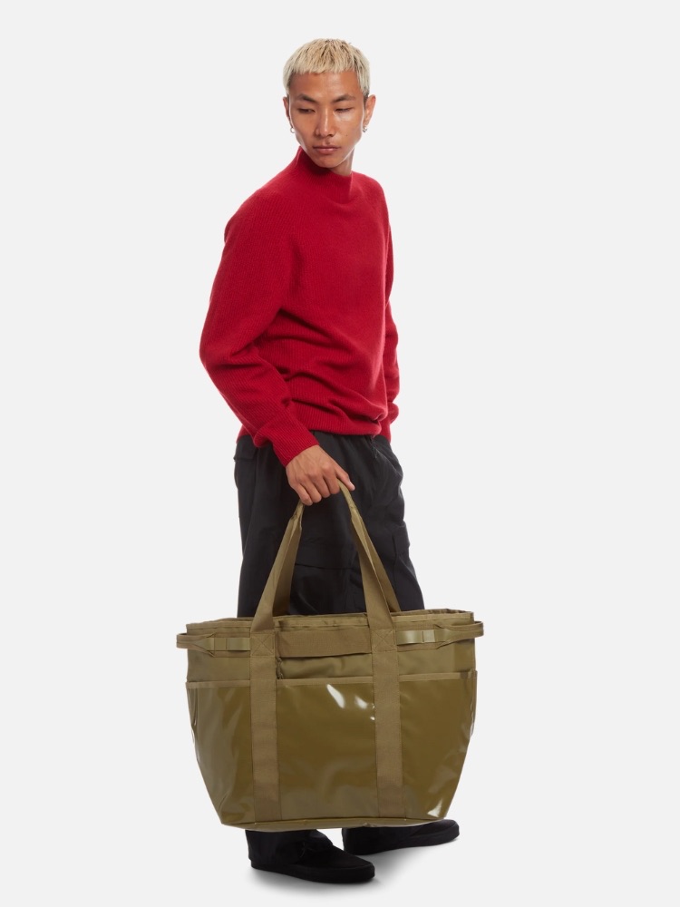 A man in a red sweater and black pants holding a large olive green tote bag, standing against a white background.