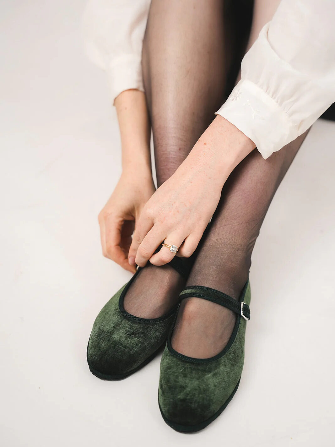 A person sitting with crossed legs, wearing dark green mary jane shoes and sheer black tights, adjusts a shoe with her hands.