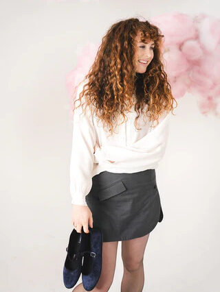 A woman with curly hair smiles, holding a pair of shoes, standing against a soft pink cloud background. she wears a white shirt and black skirt.