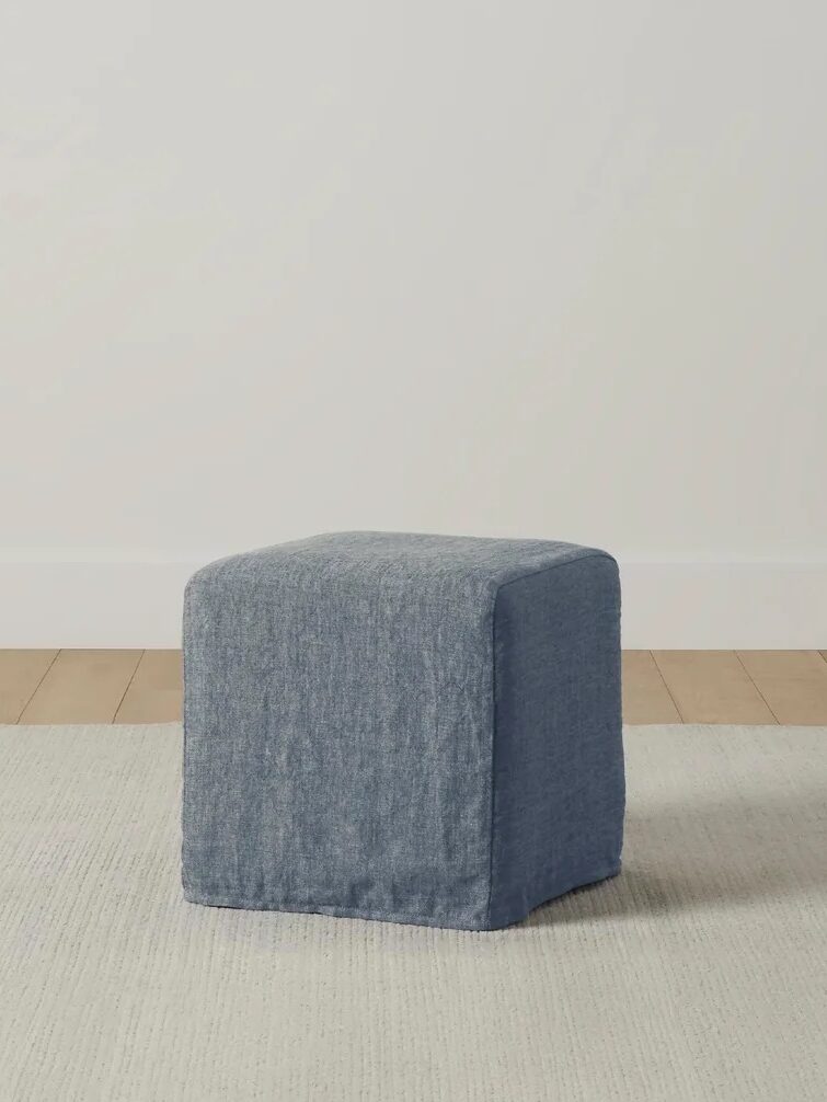 A simple blue fabric ottoman on a beige rug in a room with wooden flooring and a light gray wall.