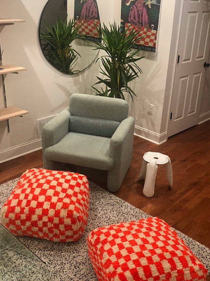 A cozy corner with a blue armchair, two red and white checkered poufs, surrounded by potted plants, a rug, and a circular mirror on the wall.
