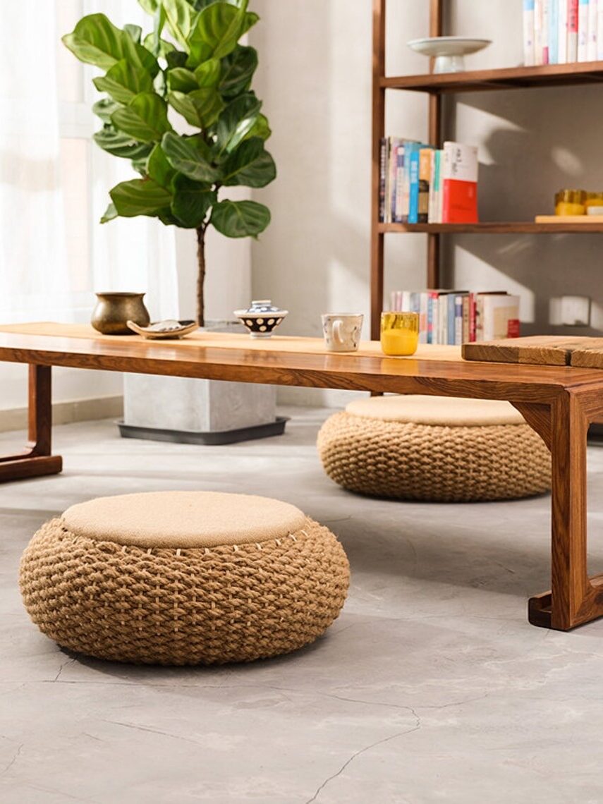 Modern minimalist living room with a wooden table, three woven ottomans, and a potted plant next to a shelving unit.