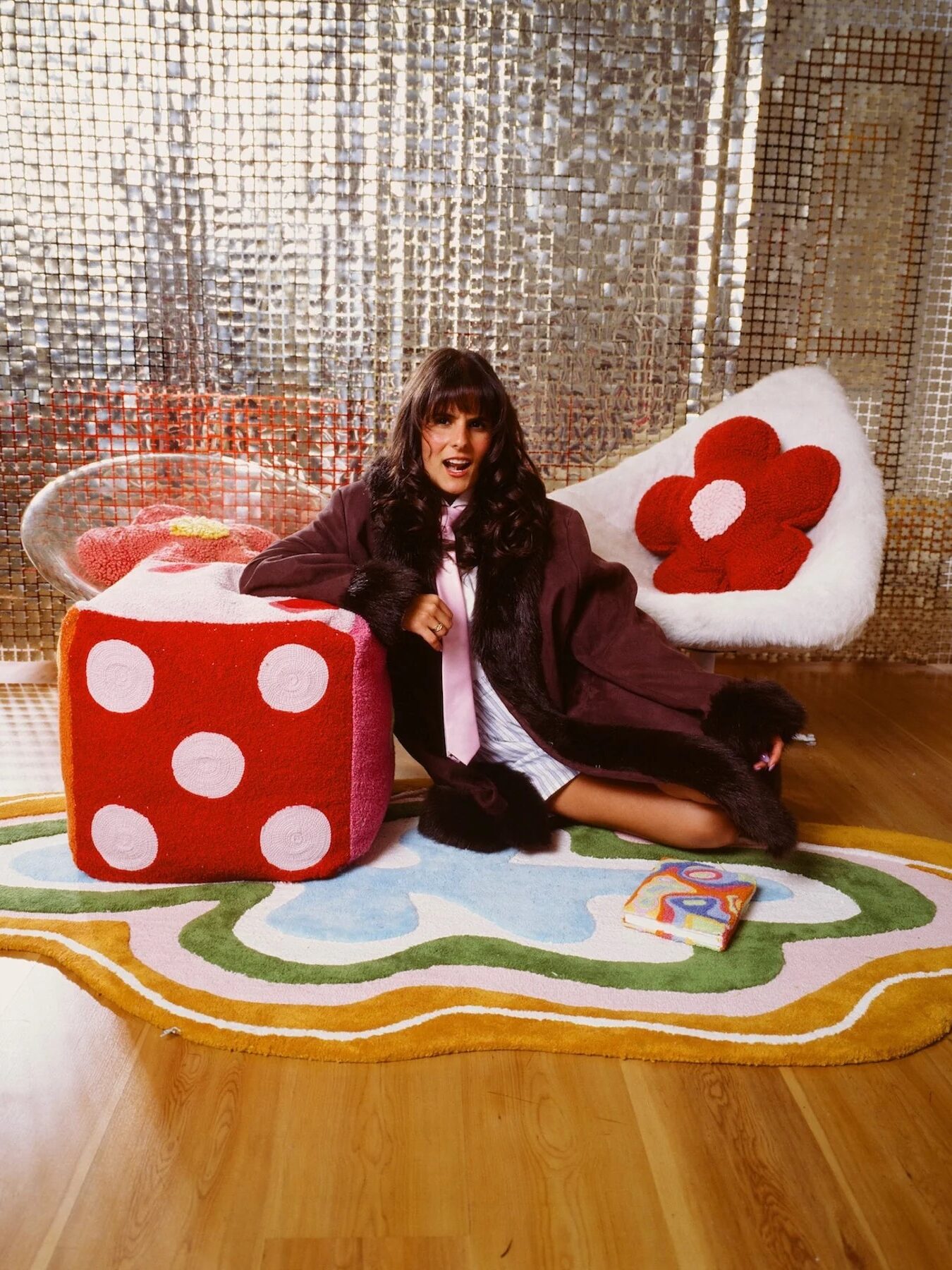 Woman in a chic coat seated on a flower-shaped rug near a large dice and a reflective backdrop, holding a phone and smiling.