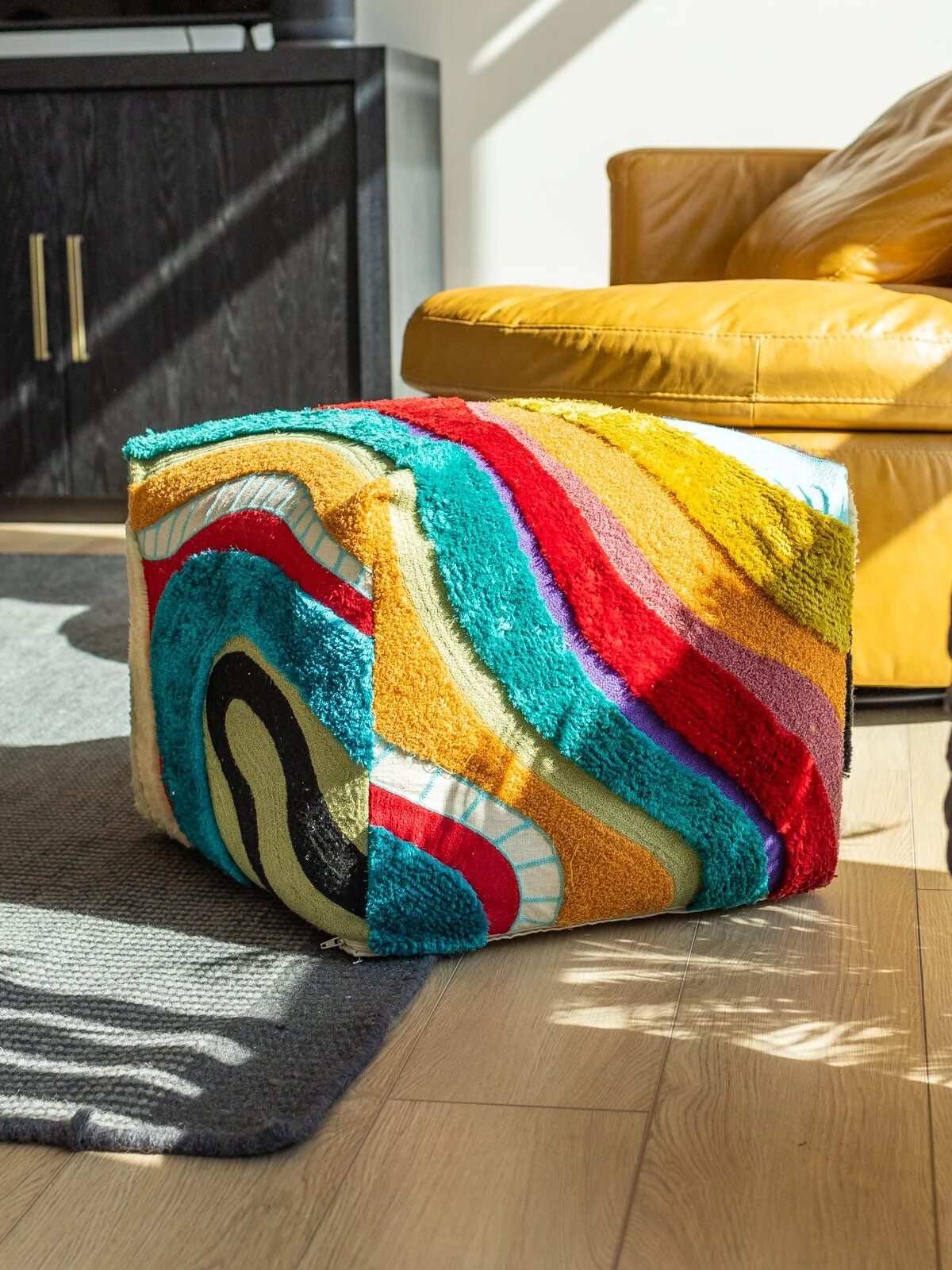 Brightly colored pouf with abstract design in a room with a hardwood floor, bathed in natural sunlight.