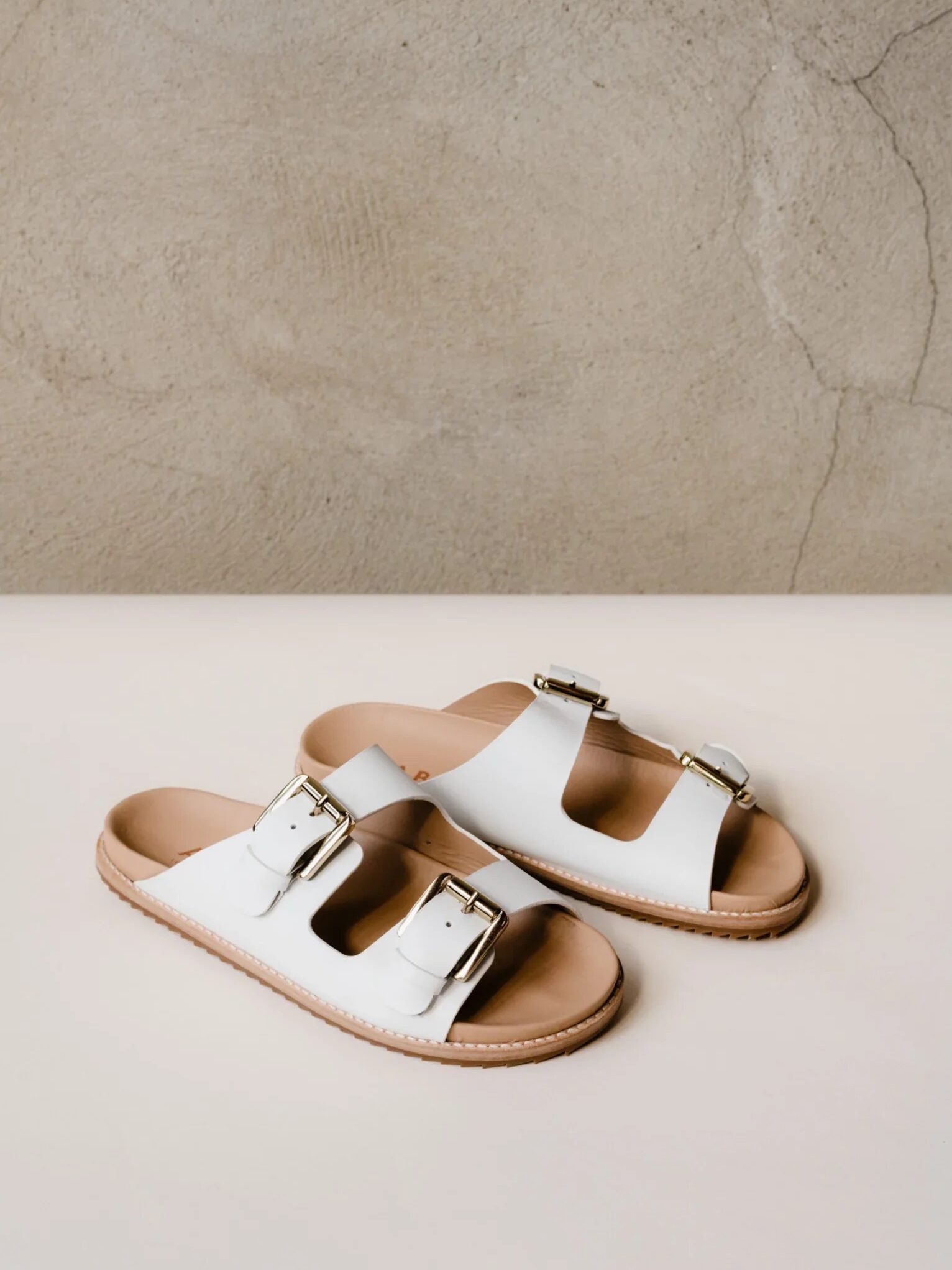 A pair of white sandals with buckle details on a beige surface against a textured grey wall.