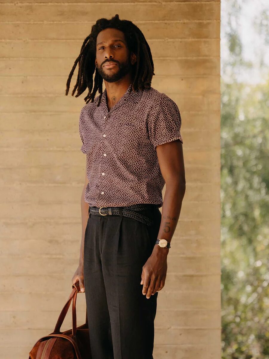 A man standing beside a concrete wall wearing a patterned shirt and black trousers, holding a brown leather bag.