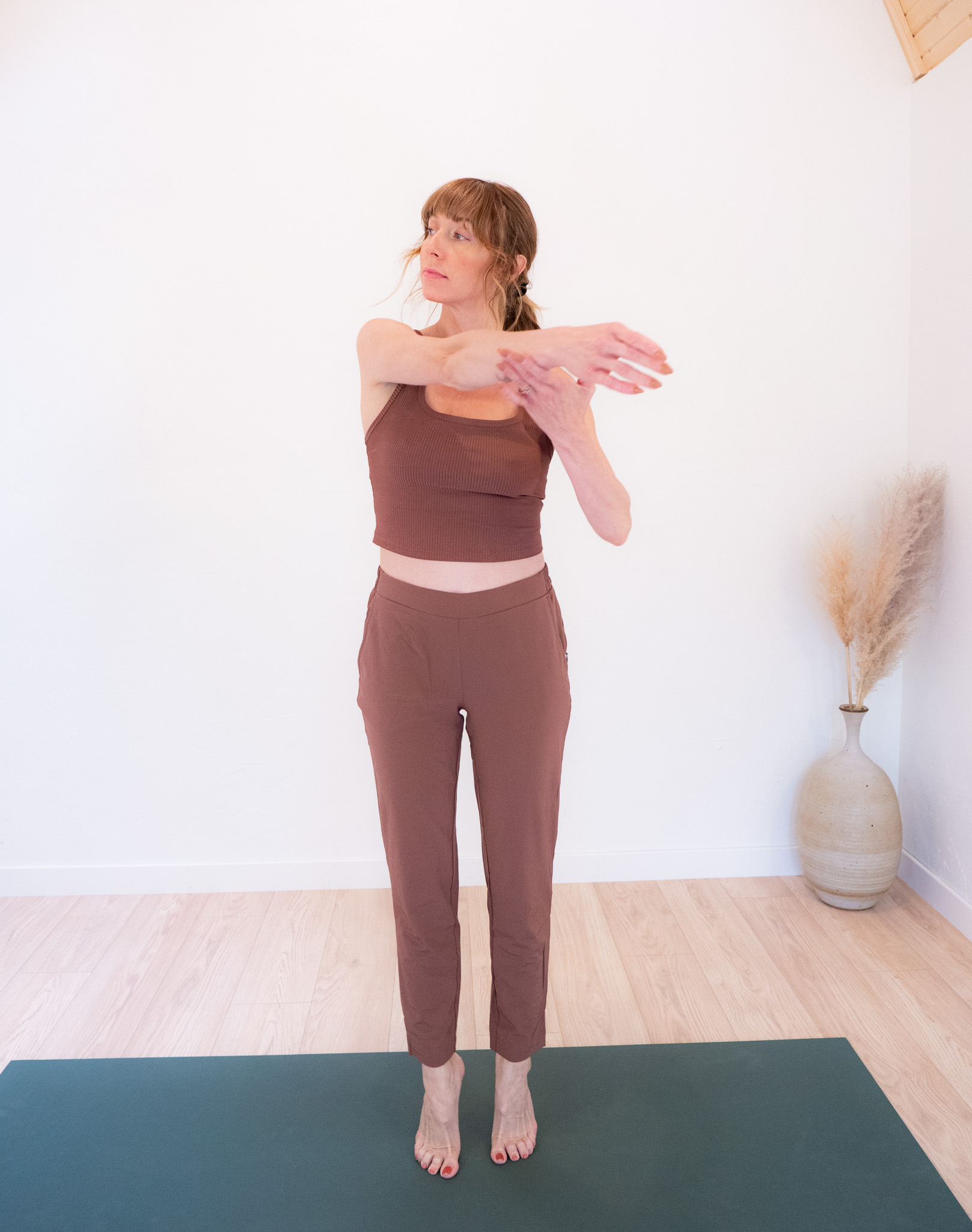 Woman in burgundy athletic wear stretching arms across her chest in a bright yoga studio.