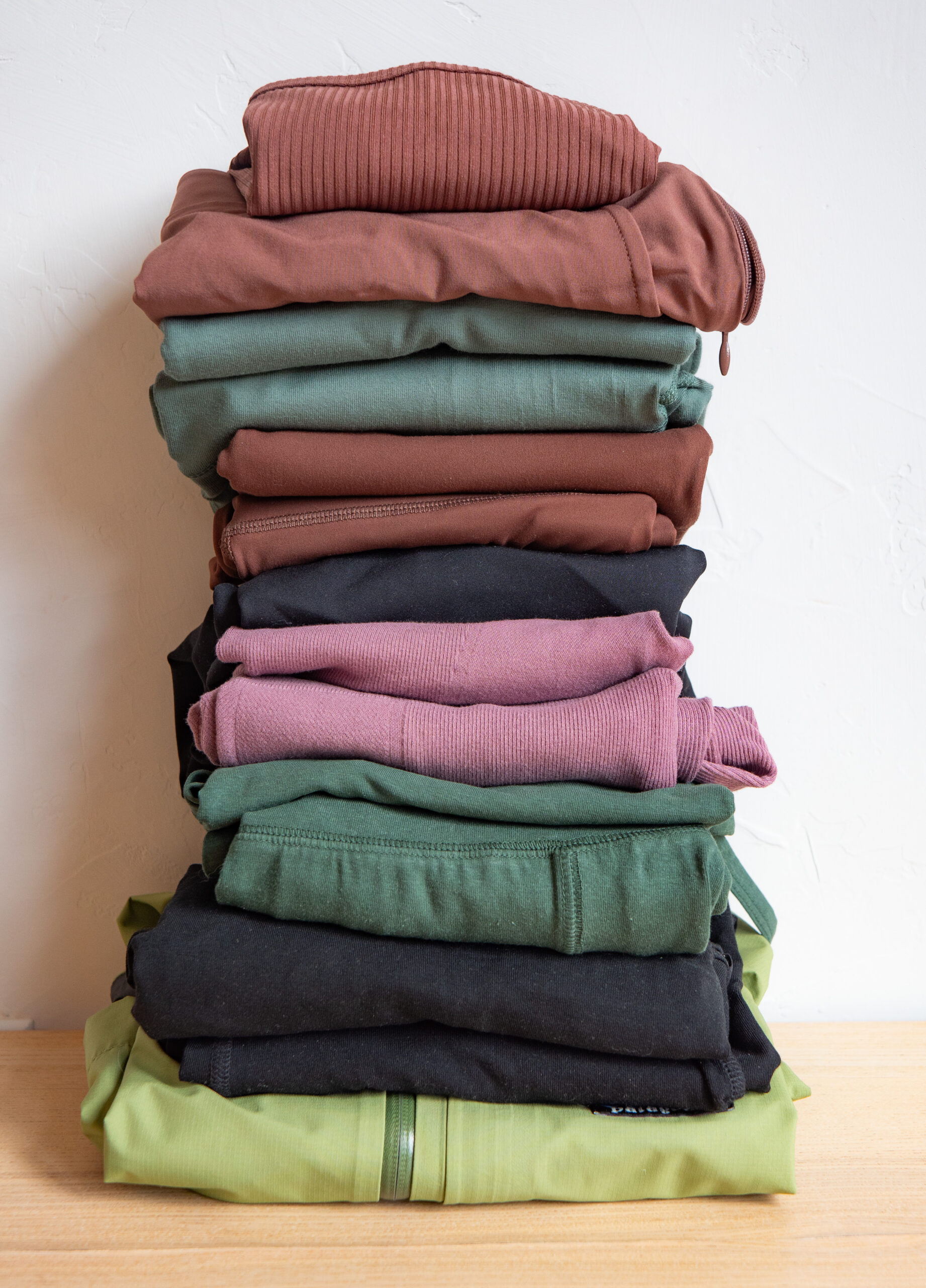 A neat stack of folded clothes in various colors including green, black, pink, and brown against a plain background.