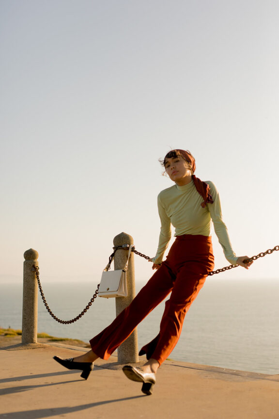 A woman in stylish attire poses confidently next to a rail by the sea, bathed in golden sunlight.