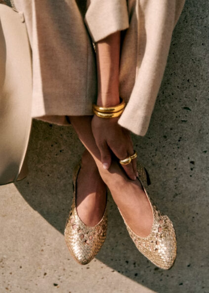 A close-up of a woman's feet in gold textured flats, her fingers adorned with rings and bracelets, under the hem of a beige coat.