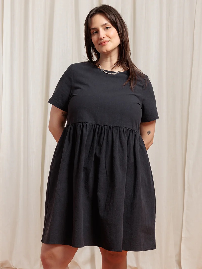 A woman in a simple black dress stands confidently, hands on hips, against a neutral-toned curtain background.