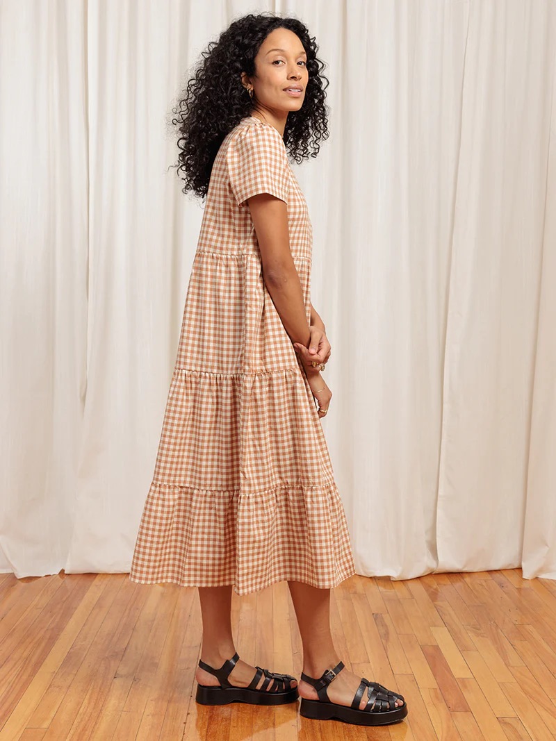 A woman with curly hair wearing a checkered midi dress and black sandals, standing in front of a plain white backdrop, looking over her shoulder.