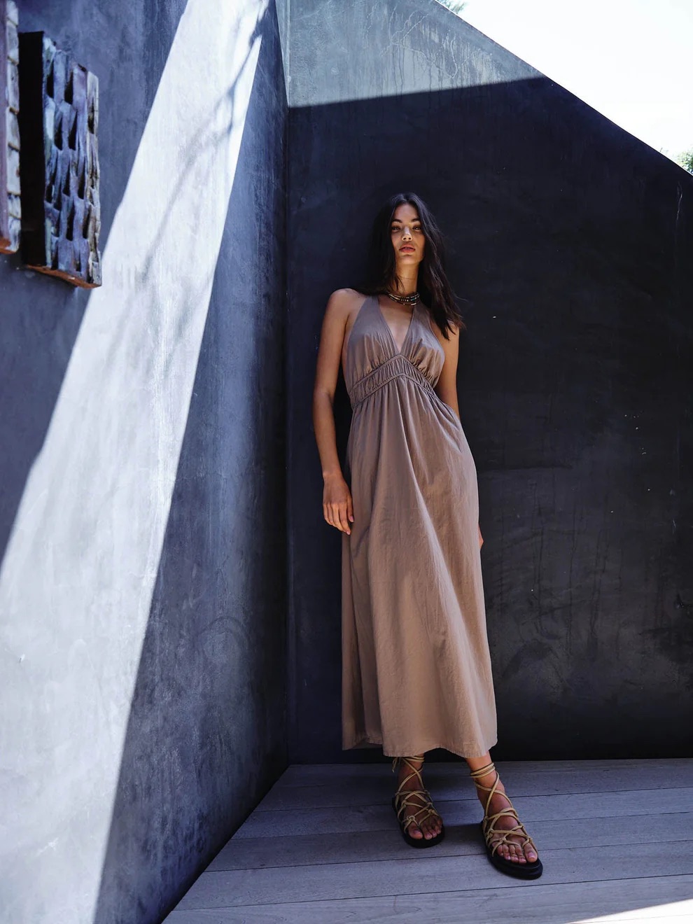 A woman in a beige dress stands confidently between dark architectural walls, with light casting shadows around her.