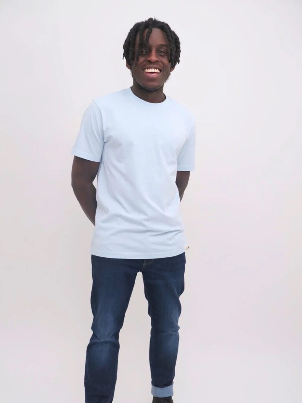 A young man in a white t-shirt and blue jeans stands smiling against a light background.