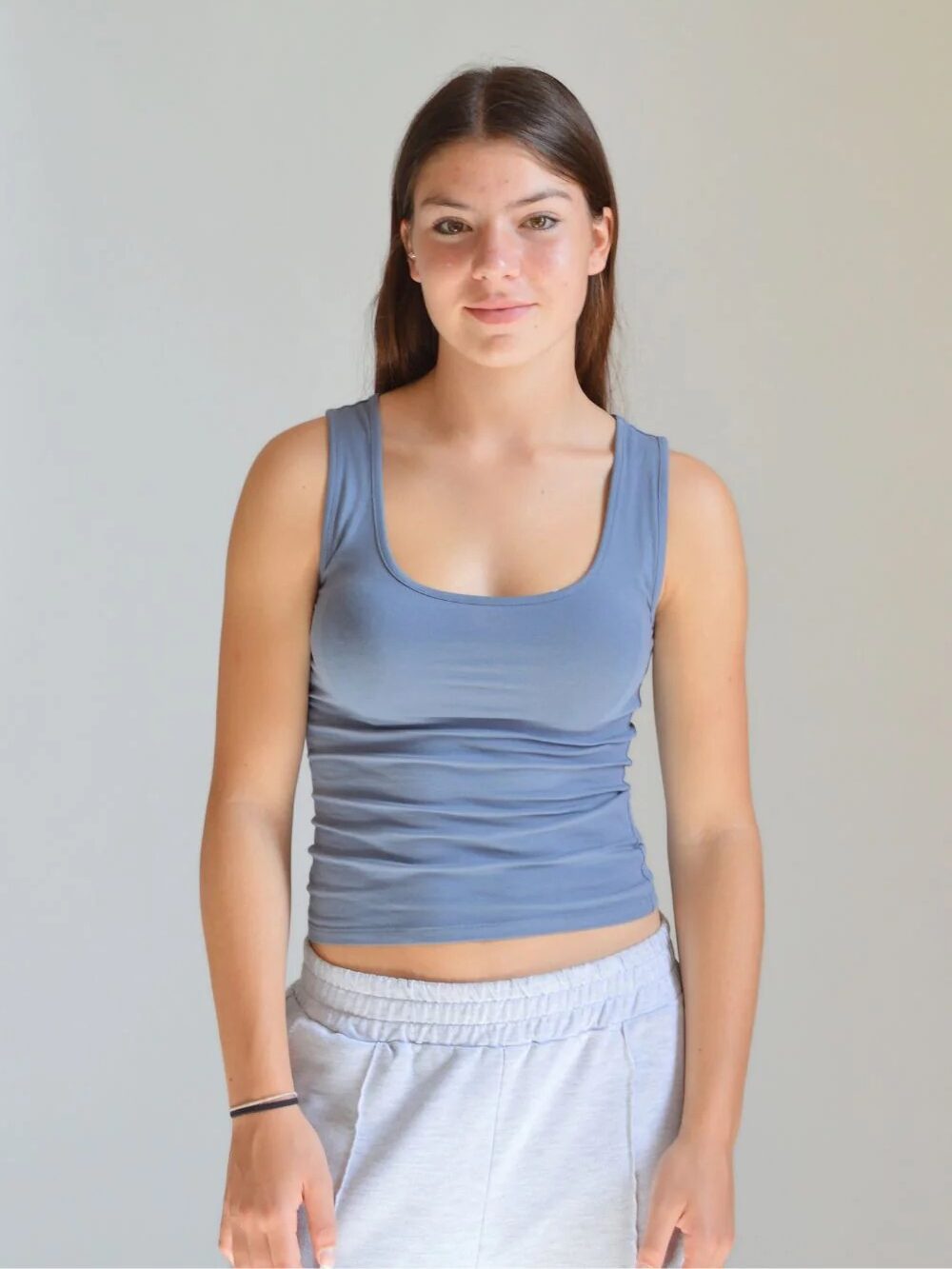 Young woman in a blue tank top and grey sweatpants standing against a light grey background.