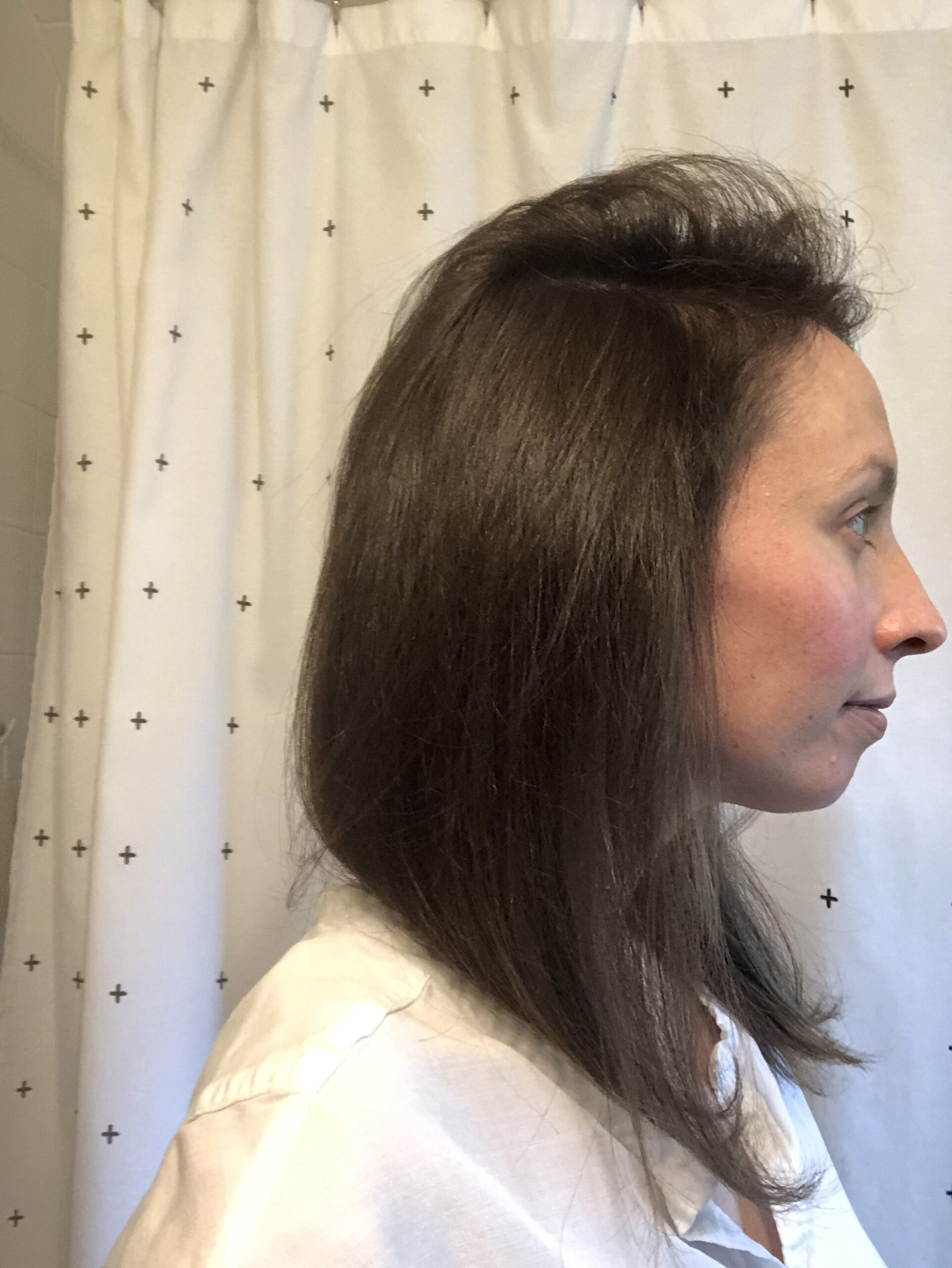 Profile view of a woman with medium-length brown hair, wearing a white shirt, standing against a curtain with a cross pattern.