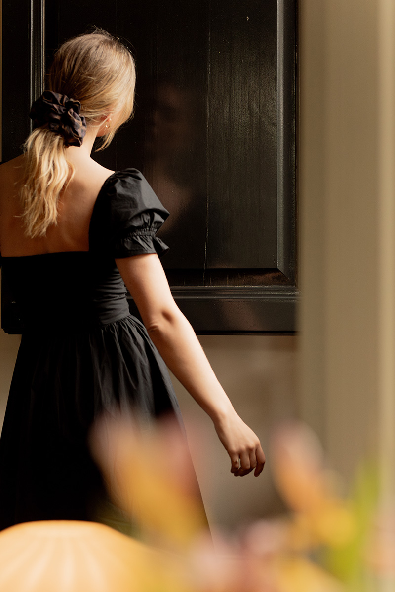 Woman in a black dress standing by a window, viewed from behind, with a focus on her arm and a blurred foreground of autumn leaves.
