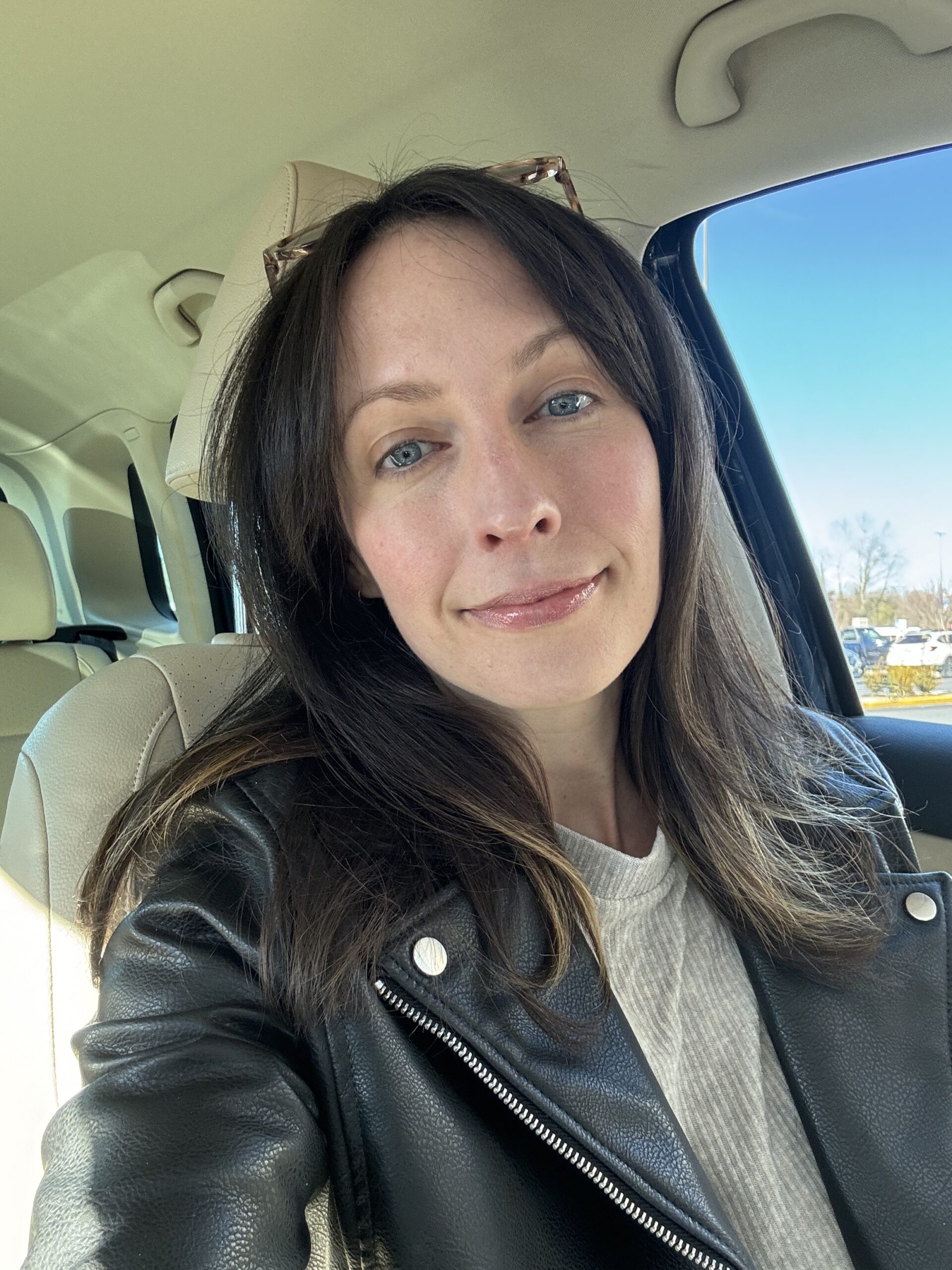 A woman with medium-length brown hair and blue eyes, wearing a white top and black leather jacket, smiling subtly in a car.