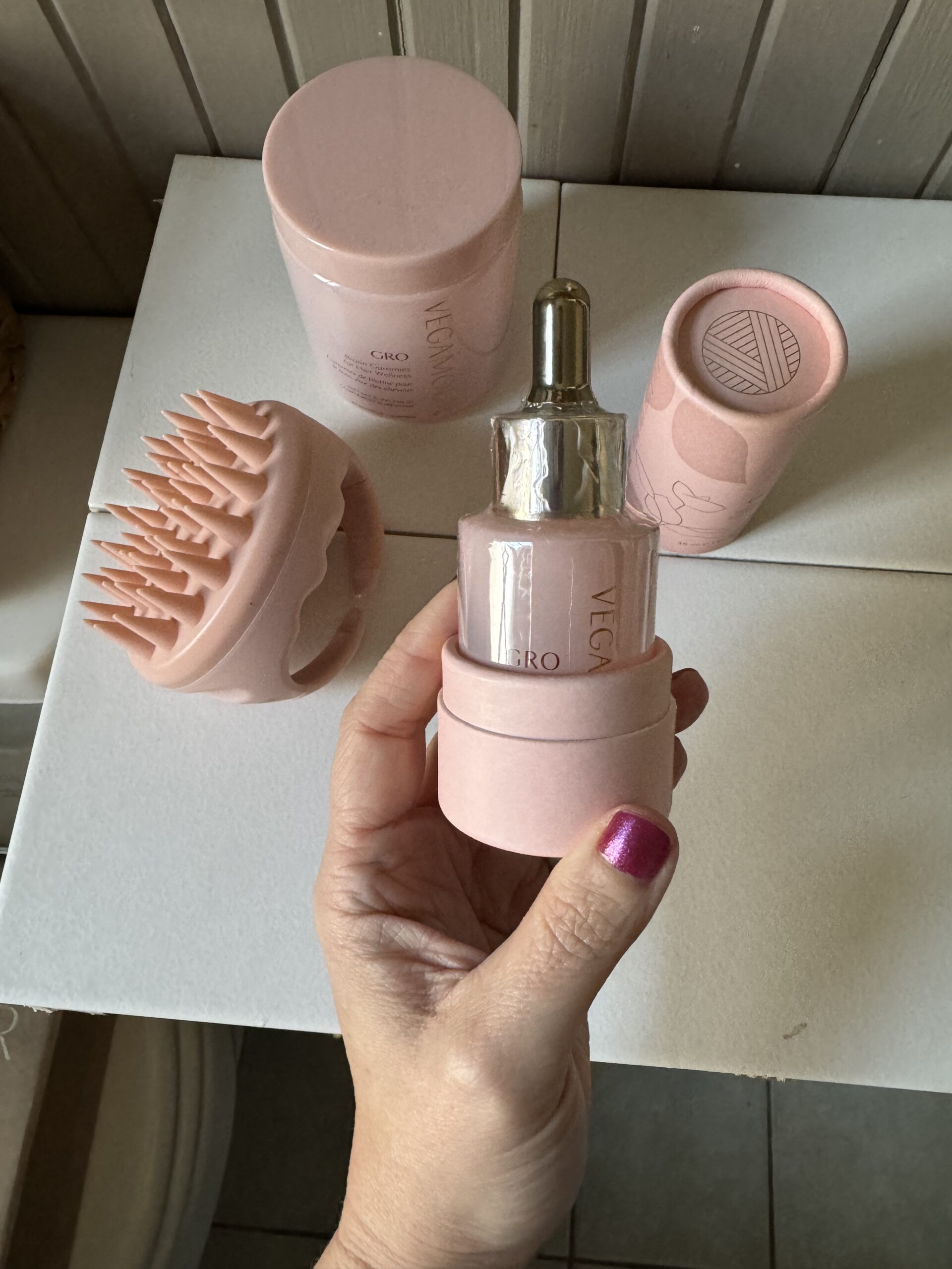 A hand holding a bottle with a dropper, surrounded by a brush, a squeezer, and other pink skincare products on a table.