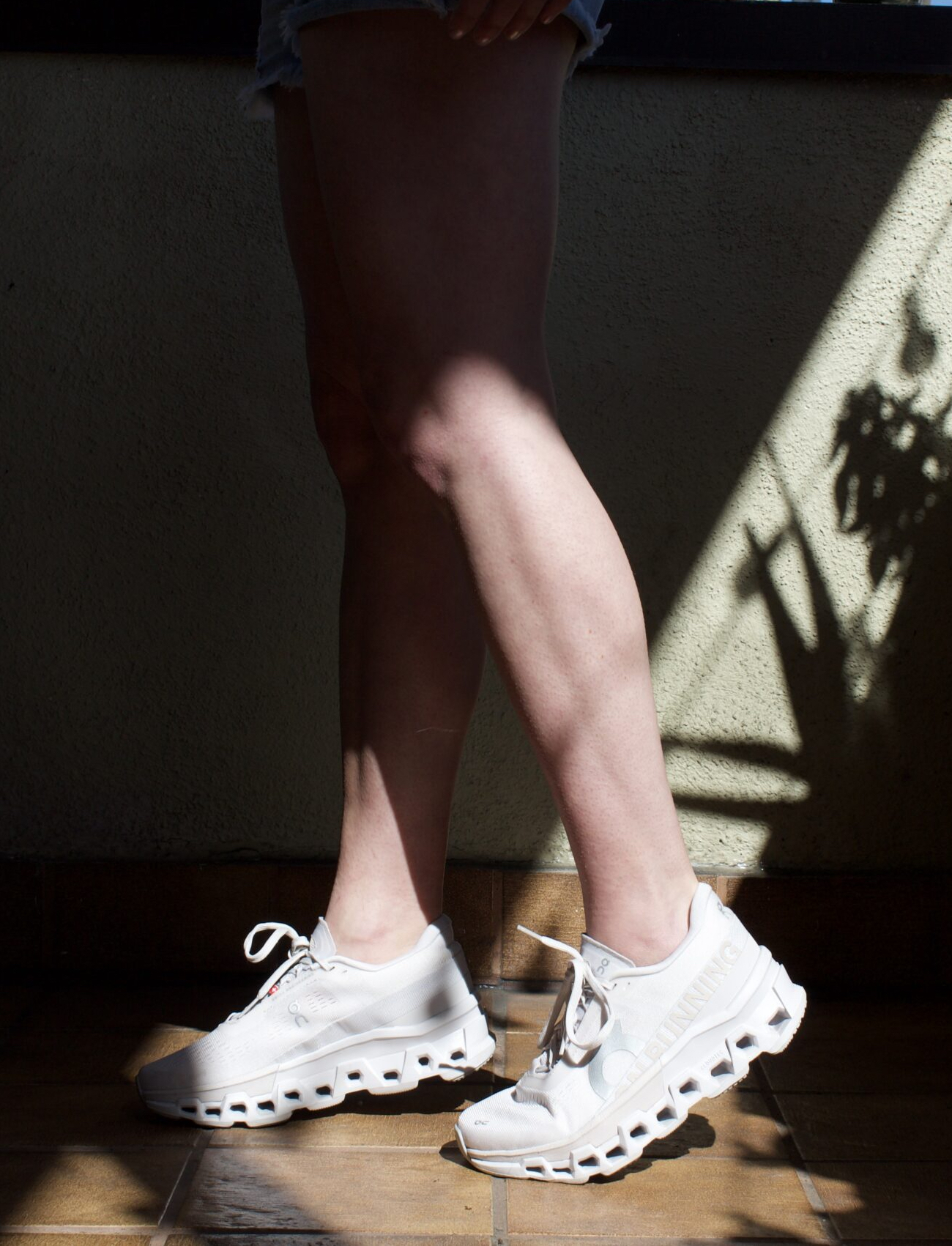 A person standing in sunlight, casting sharp shadows, wearing white sneakers and denim shorts, focused on legs.