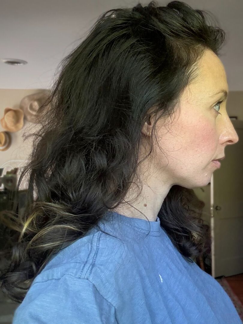 Side profile of a woman with shoulder-length dark hair in a blue shirt, standing indoors near a bookshelf.