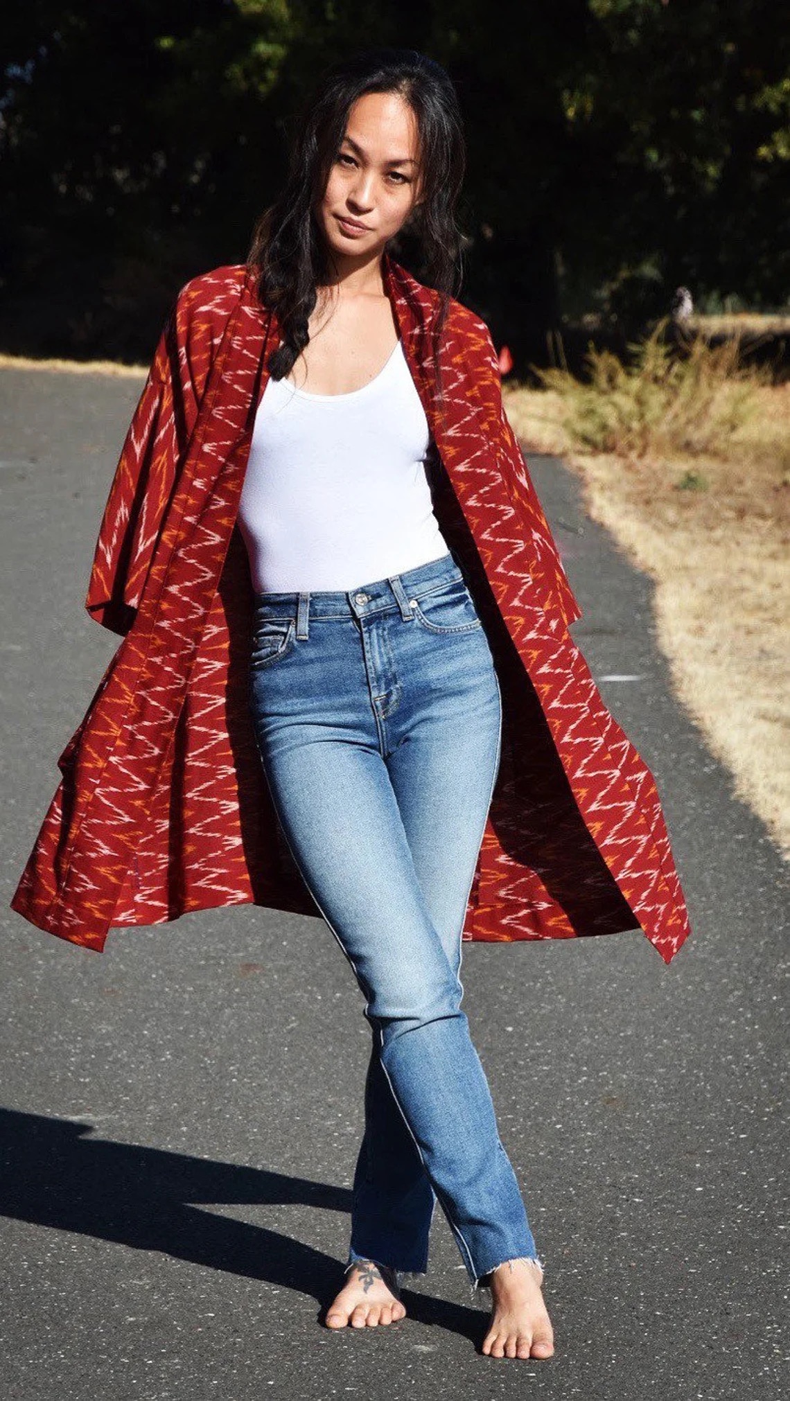 Woman walking on a road wearing jeans and a white top, with a red patterned cardigan flowing behind her.